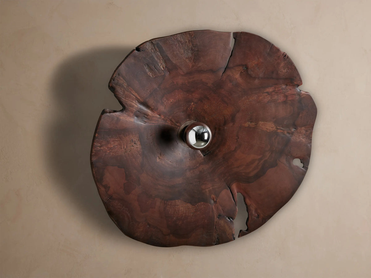 Arruga Sconce: Domed, irregular dark milpa wood sconce with natural grain & cracks, a silver bulb glowing from its center on a wall.