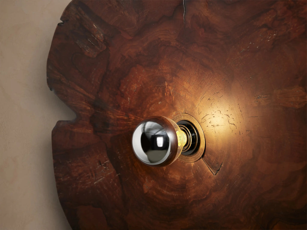 Arruga Sconce: Irregular, dark brown milpa wood wall sconce with natural grain & cracks, a central metallic bulb, on a tan wall.