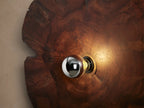 Arruga Sconce: Irregular, dark brown milpa wood wall sconce with natural grain & cracks, a central metallic bulb, on a tan wall.