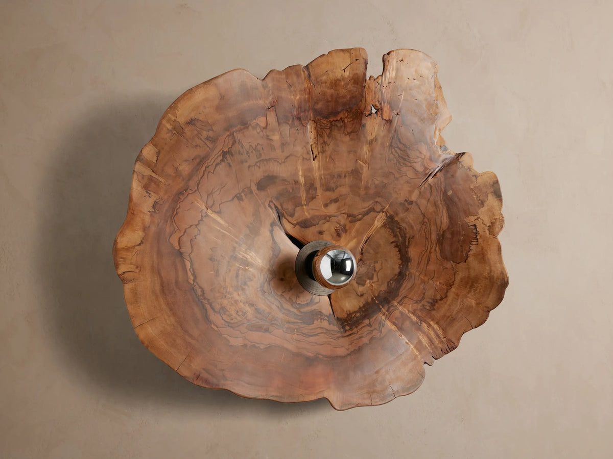 Arruga Sconce: Irregular milpa wood slice with warm brown grain & a central metallic-dipped bulb glowing on a textured brown wall.