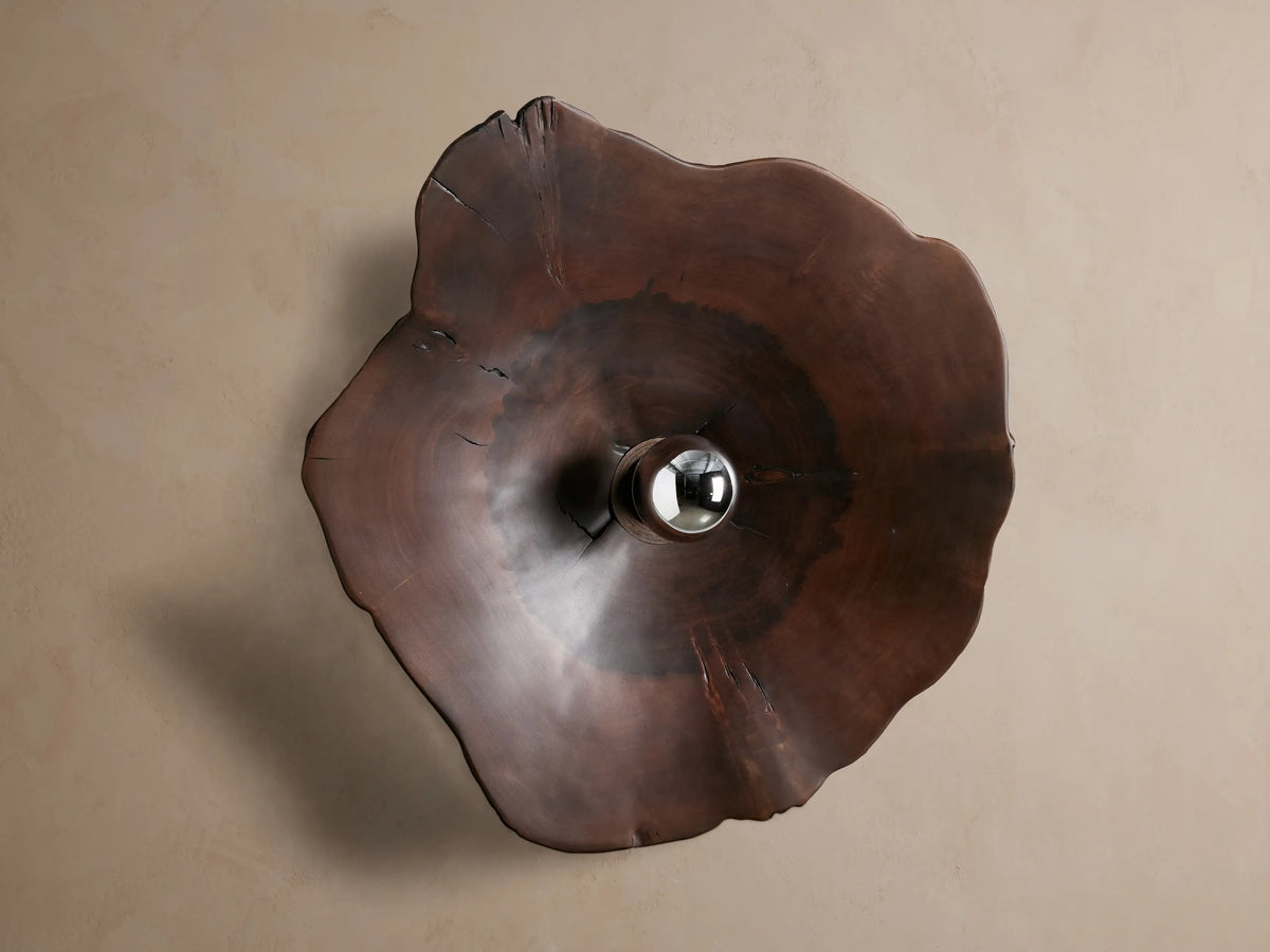 Dark brown, irregular milpa wood Arruga Sconce with a metallic-dipped bulb glowing from its dome center, on a beige wall.