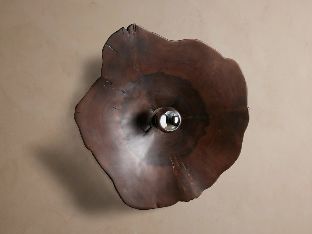 Dark brown, irregular milpa wood Arruga Sconce with a metallic-dipped bulb glowing from its dome center, on a beige wall.