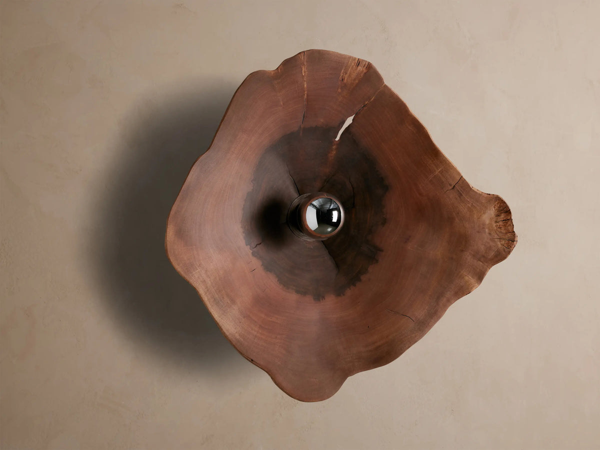 Arruga Sconce: Organic milpa wood sconce, dark brown with natural cracks, featuring a metallic silver bulb glowing golden on a taupe wall.