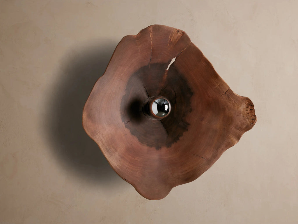 Arruga Sconce: Organic milpa wood sconce, dark brown with natural cracks, featuring a metallic silver bulb glowing golden on a taupe wall.