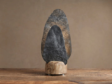 Shinto Table Lamp. Two irregular slate stone shades, dark gray front, lighter illuminated rear, in a natural wood base on a wood table.