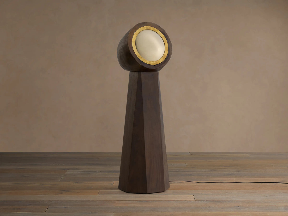 Agustin Floor Lamp: Dark wood faceted base with a round, gold-rimmed opaque glass light fixture, glowing warmly.