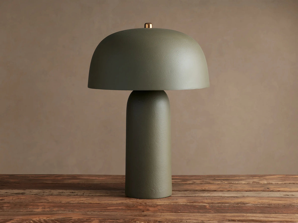 Ira Table Lamp: Sage green lamp with a dome shade, cylindrical base, and small brass accent, emitting a warm glow on wood.