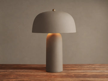 Ira Table Lamp, a textured taupe mushroom-shaped lamp with a gold accent, casting a warm glow on a wooden table.