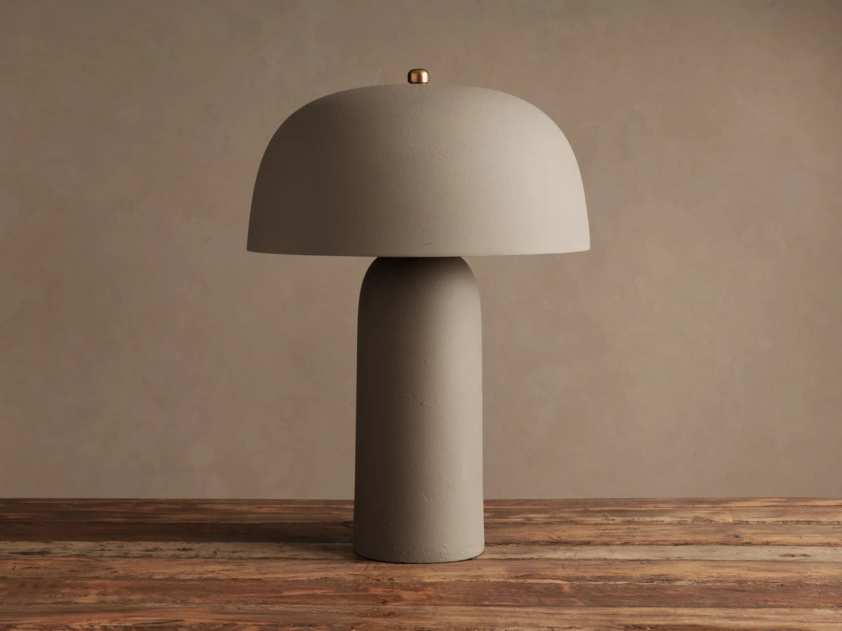 Ira Table Lamp, a textured taupe mushroom-shaped lamp with a gold accent, casting a warm glow on a wooden table.