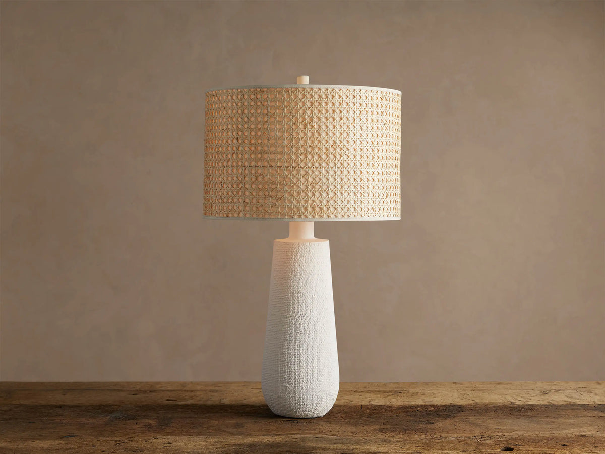 Stucco Table Lamp with a textured white conical base and a natural woven cane cylindrical shade, on a rustic wooden table.