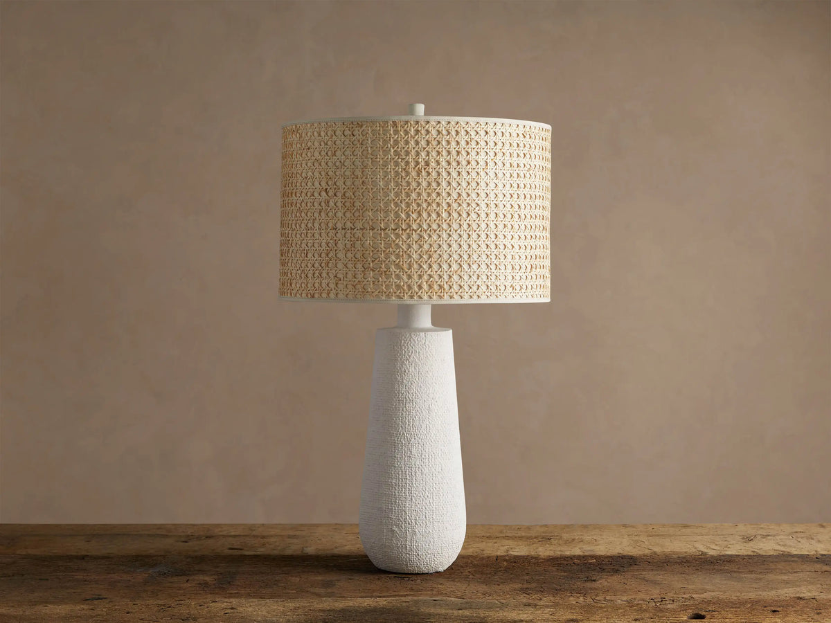 Stucco Table Lamp with a textured white conical base and a natural woven cane cylindrical shade, on a rustic wooden table.