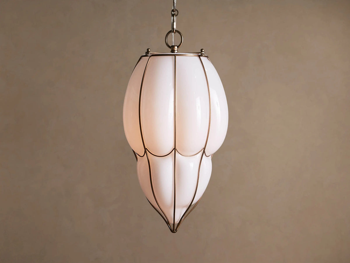 Margolis Large Pendant: Elongated white opaque glass pendant light with brass-finished frame, glowing softly, against a neutral wall.