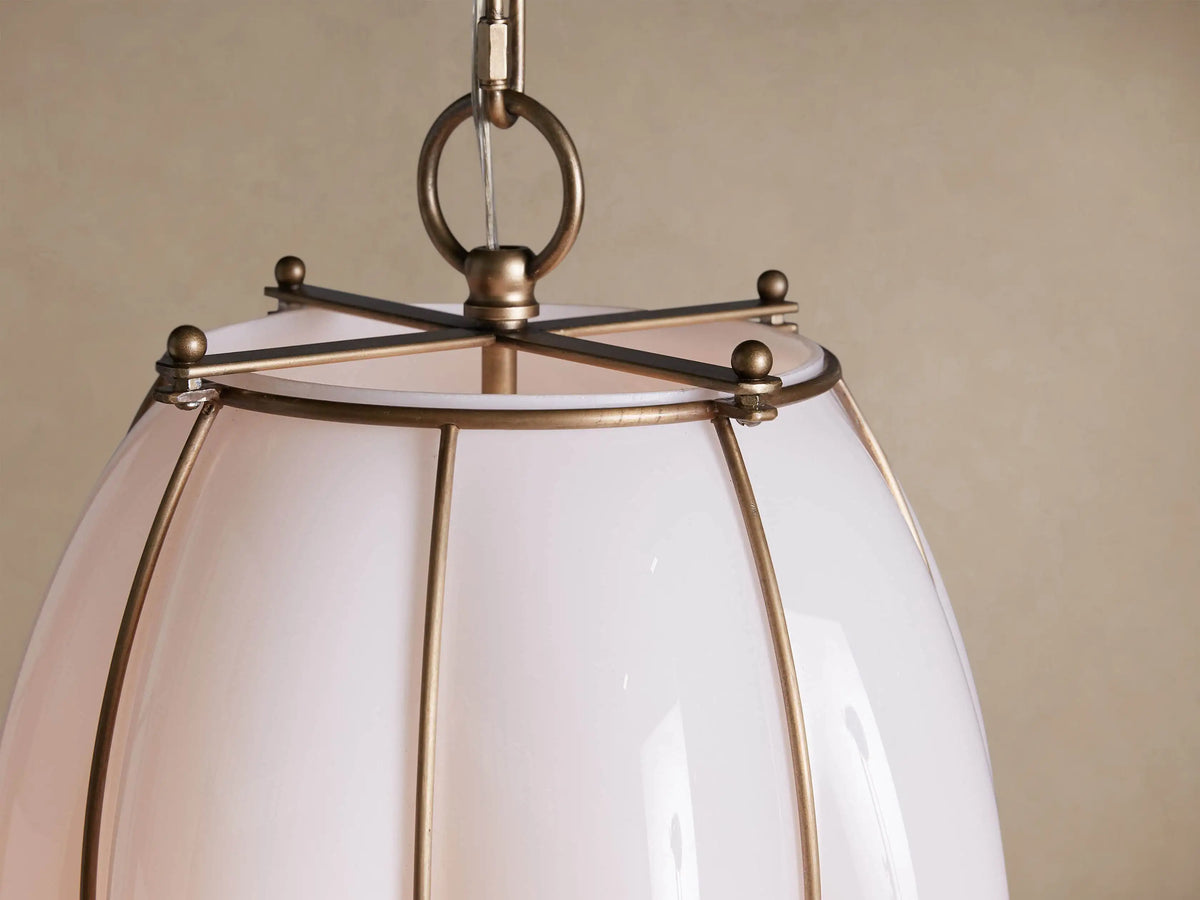 Margolis Large Pendant: Elongated white opaque glass pendant light with brass-finished frame, glowing softly, against a neutral wall.