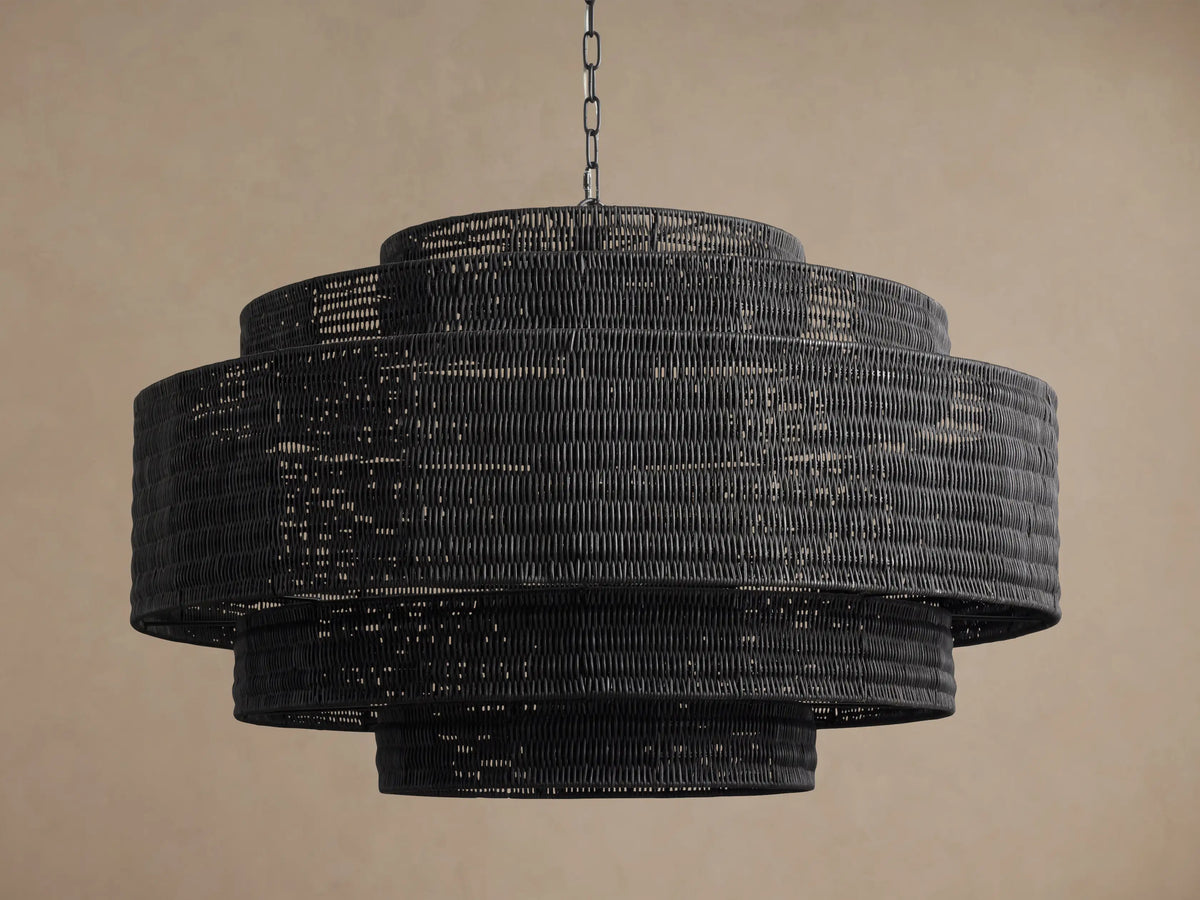Paloma Chandelier, large tiered cylindrical black handwoven rattan fixture hanging from a silver chain against a brown wall.