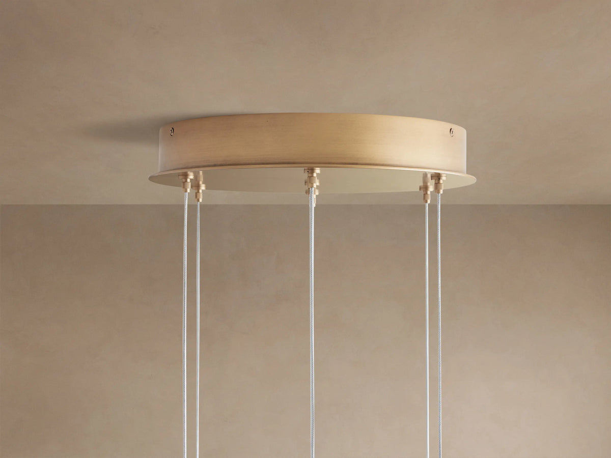 Arica Round Chandelier: brass-toned round canopy mounted on a tan ceiling, with several exposed airplane cables hanging.