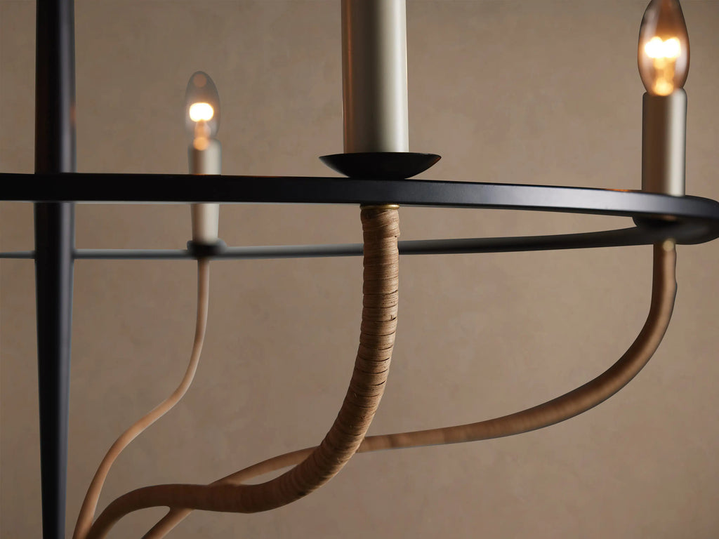 Black iron Rosalind Rattan Chandelier with 5 candle-style bulbs, wrapped in natural rattan, suspended against a tan wall.