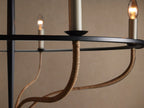 Black iron Rosalind Rattan Chandelier with 5 candle-style bulbs, wrapped in natural rattan, suspended against a tan wall.