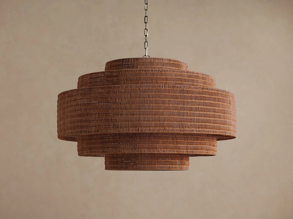 Paloma Chandelier: Tiered brown handwoven rattan chandelier with three cylindrical layers, glowing warmly from within against a neutral wall.