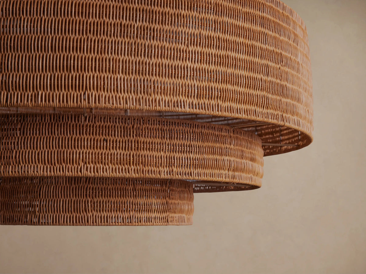 Paloma Chandelier, tiered warm brown handwoven rattan shade hanging from a dark metal chain against a plain beige wall.