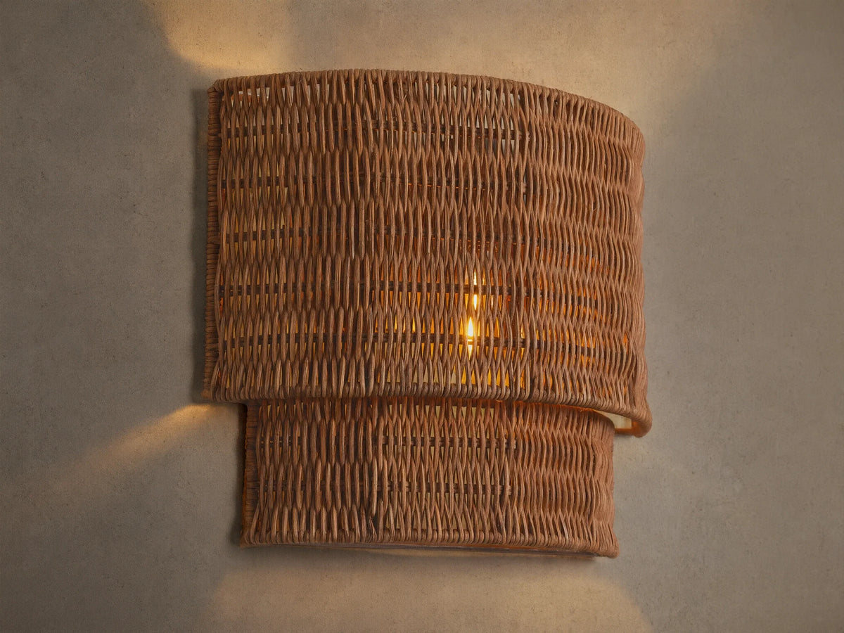 Paloma Sconce with a curved, layered handwoven rattan shade in natural brown, glowing warmly on a beige wall.