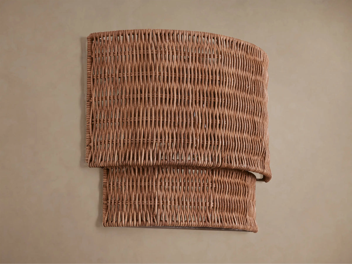 Paloma Sconce with a curved, layered handwoven rattan shade in natural brown, glowing warmly on a beige wall.