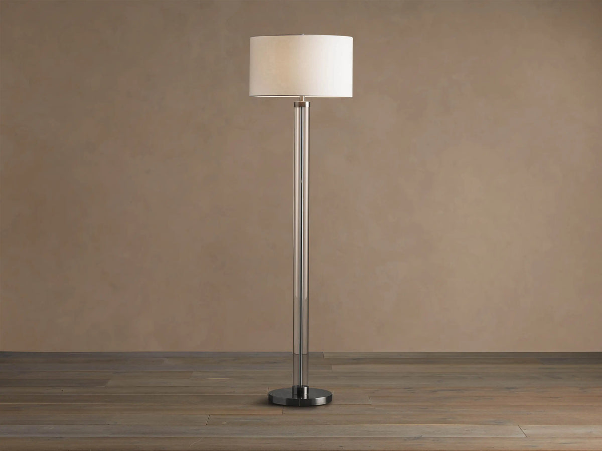 Tall Glass Column Floor Lamp with a clear glass stem, round black iron base, and a cream linen drum shade, on a wood floor.