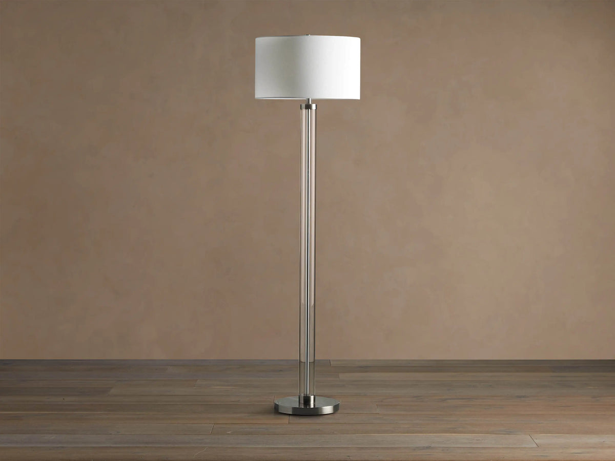 Tall Glass Column Floor Lamp with a clear glass stem, round black iron base, and a cream linen drum shade, on a wood floor.