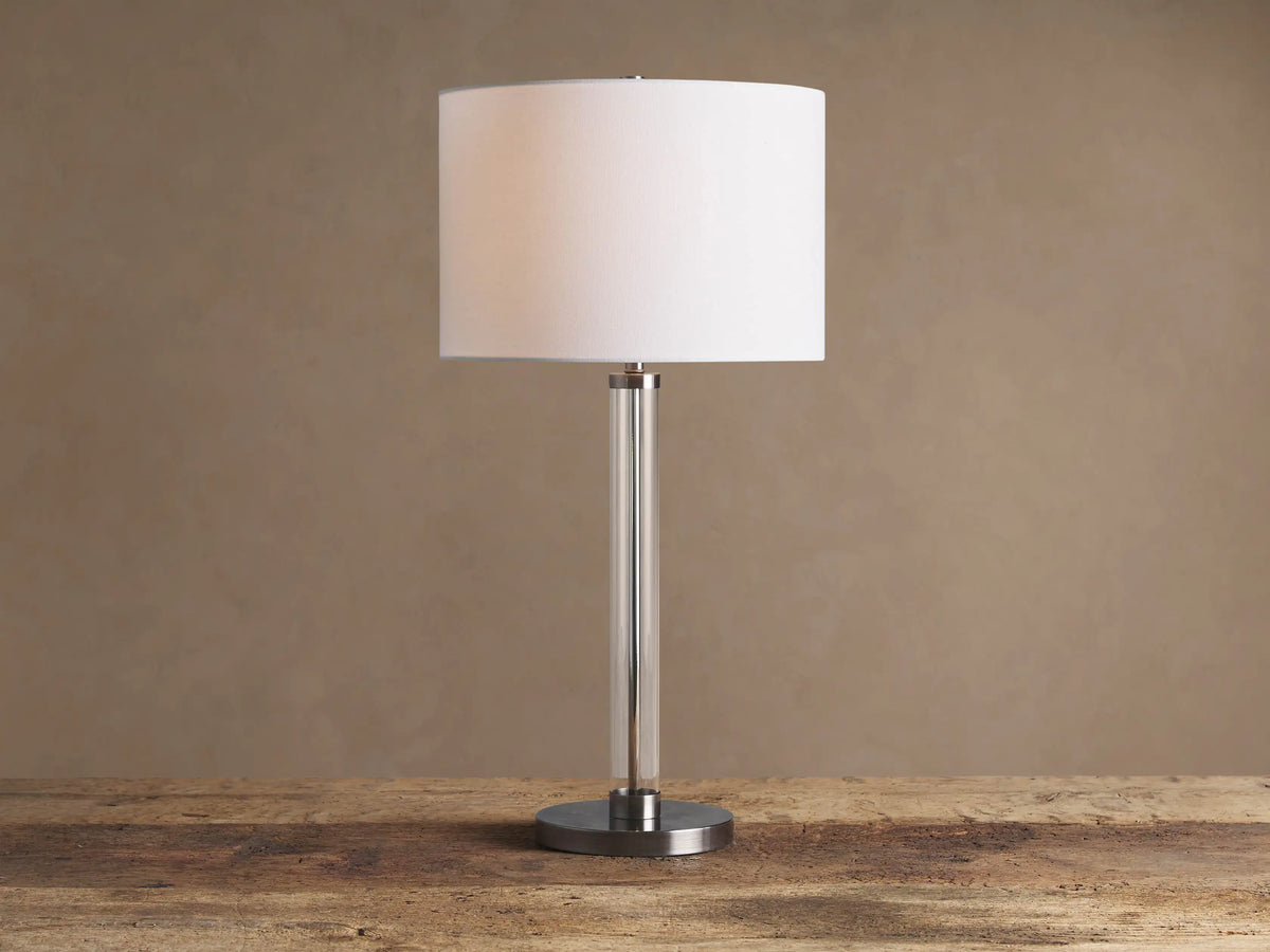 Glass Column Table Lamp with clear glass column, brushed metal base, and white linen shade on a wooden table.