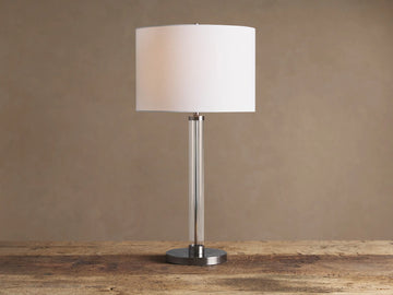 Glass Column Table Lamp with clear glass column, brushed metal base, and white linen shade on a wooden table.