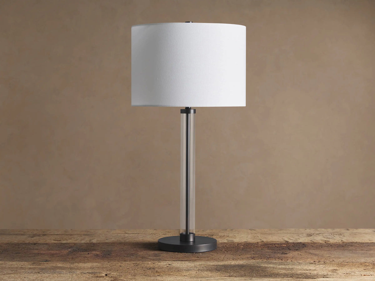 Glass Column Table Lamp with clear glass column, dark iron base, and white linen drum shade on a rustic wood table.