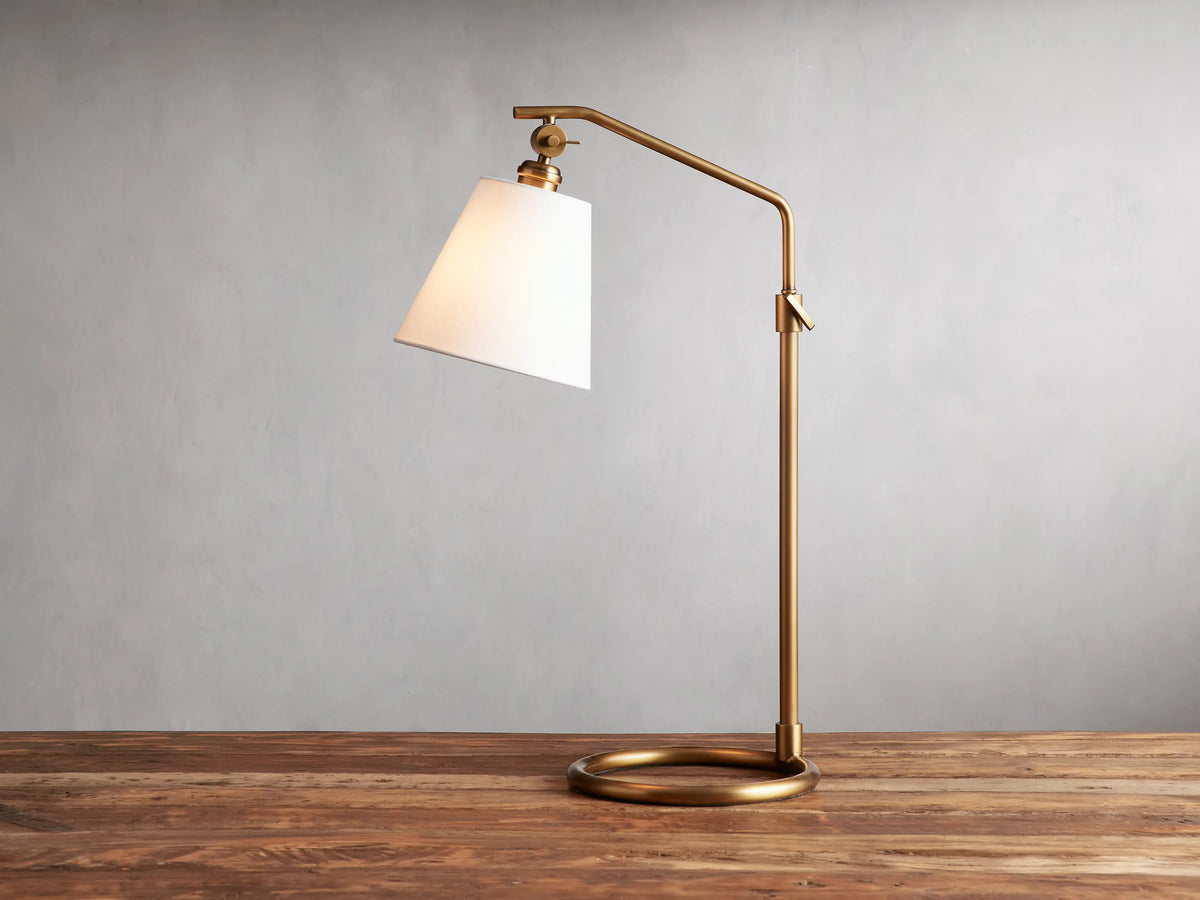 Kellen Table Lamp in antiqued-brass with white linen shade, on wood surface. Adjustable arm and round base, cord visible.