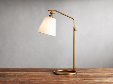 Kellen Table Lamp in antiqued-brass with white linen shade, on wood surface. Adjustable arm and round base, cord visible.