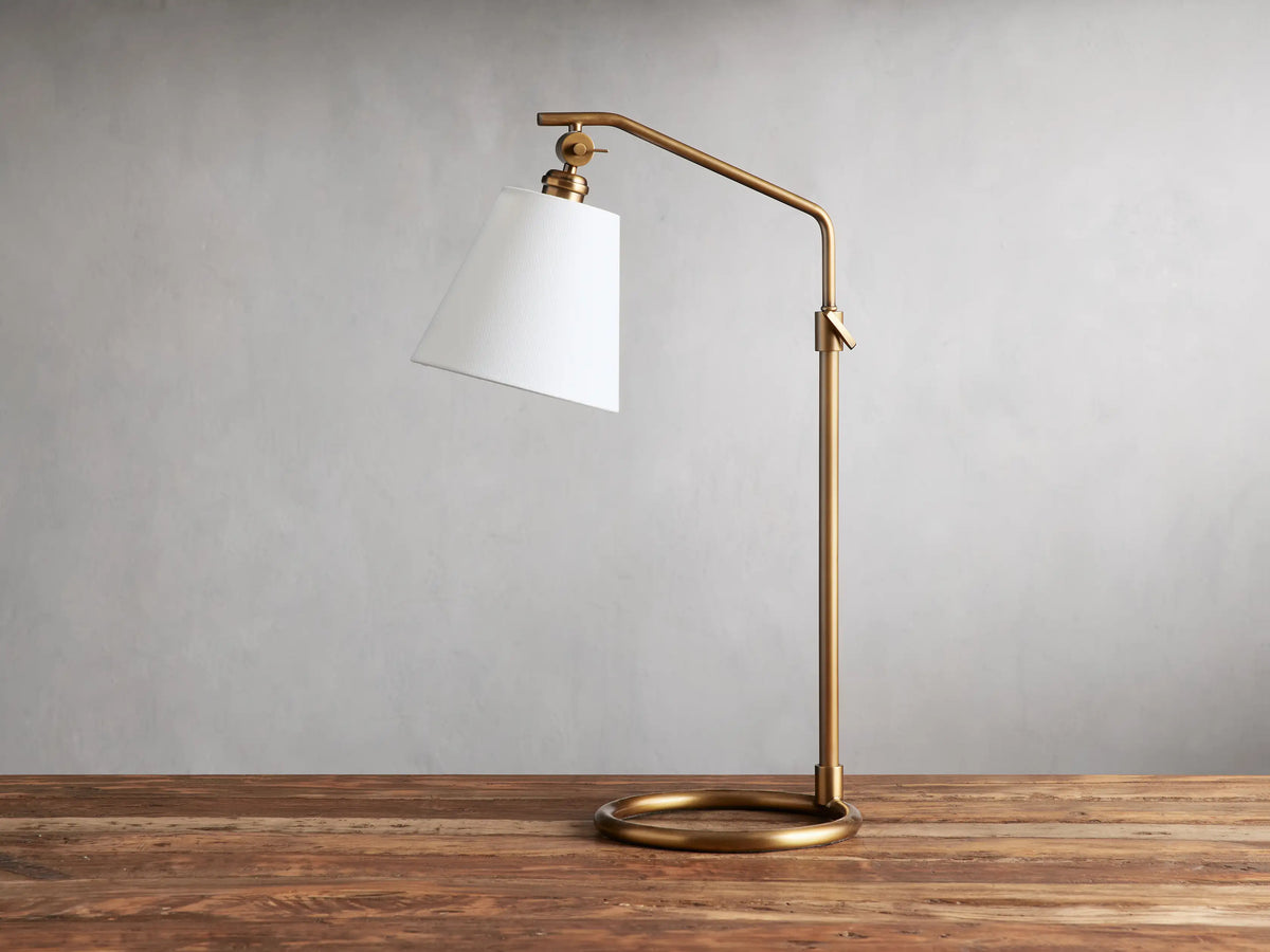 Kellen Table Lamp in antiqued-brass with white linen shade, on wood surface. Adjustable arm and round base, cord visible.