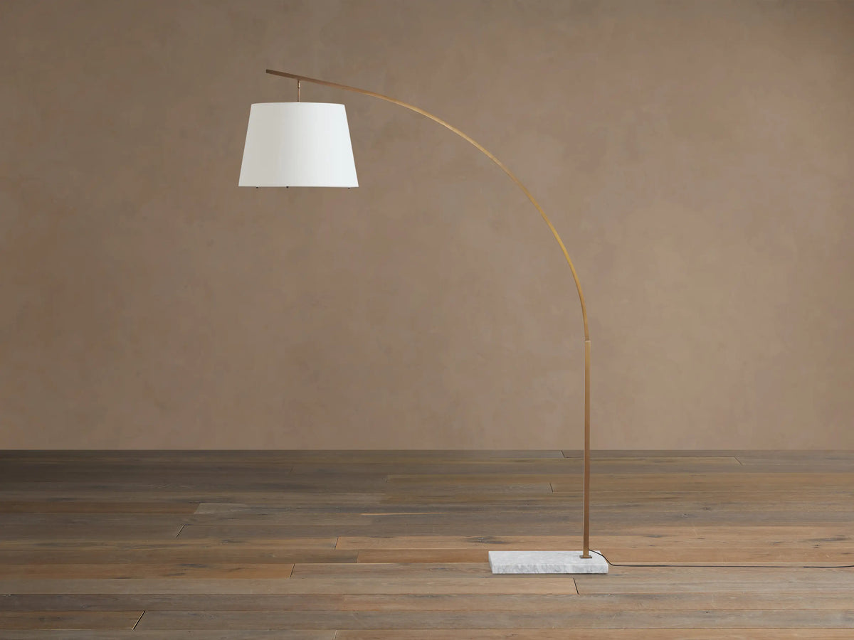 Tadeo arc floor lamp features a curved brass-colored stand, white linen drum shade, and square white marble base on a wood floor.