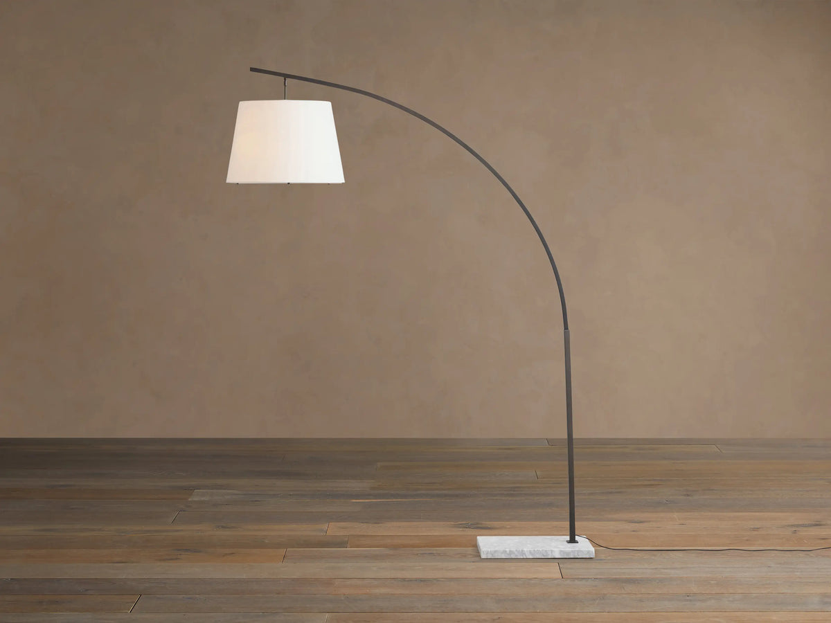 Tadeo Floor Lamp with a curved dark bronze arm, white linen shade, and rectangular white marble base on a wood floor.