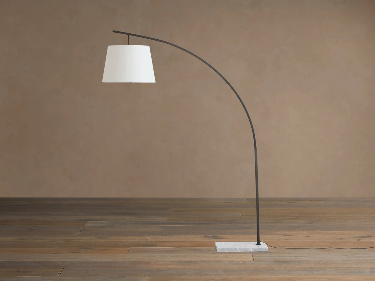 Tadeo Floor Lamp with a curved dark bronze arm, white linen shade, and rectangular white marble base on a wood floor.
