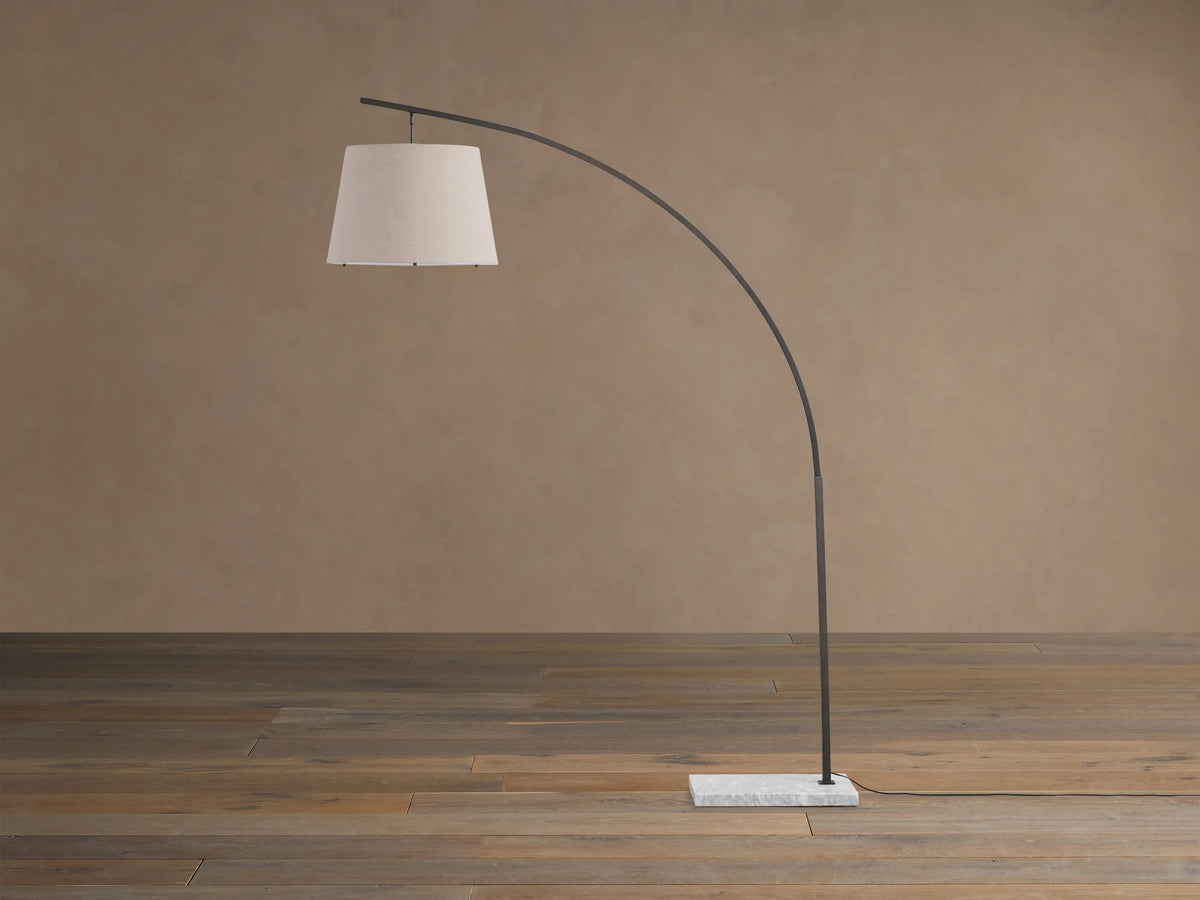 Tadeo Floor Lamp: dark arcing frame, light linen drum shade, white marble base, on wooden floor against a warm wall.