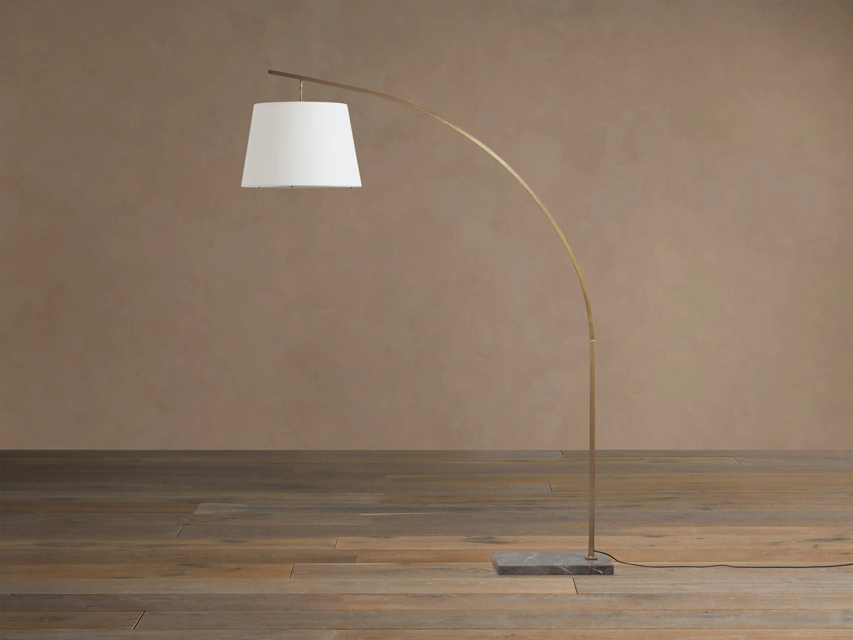 Tadeo Floor Lamp with arcing brass-toned pole, white linen shade, and dark marble base on a wooden floor.