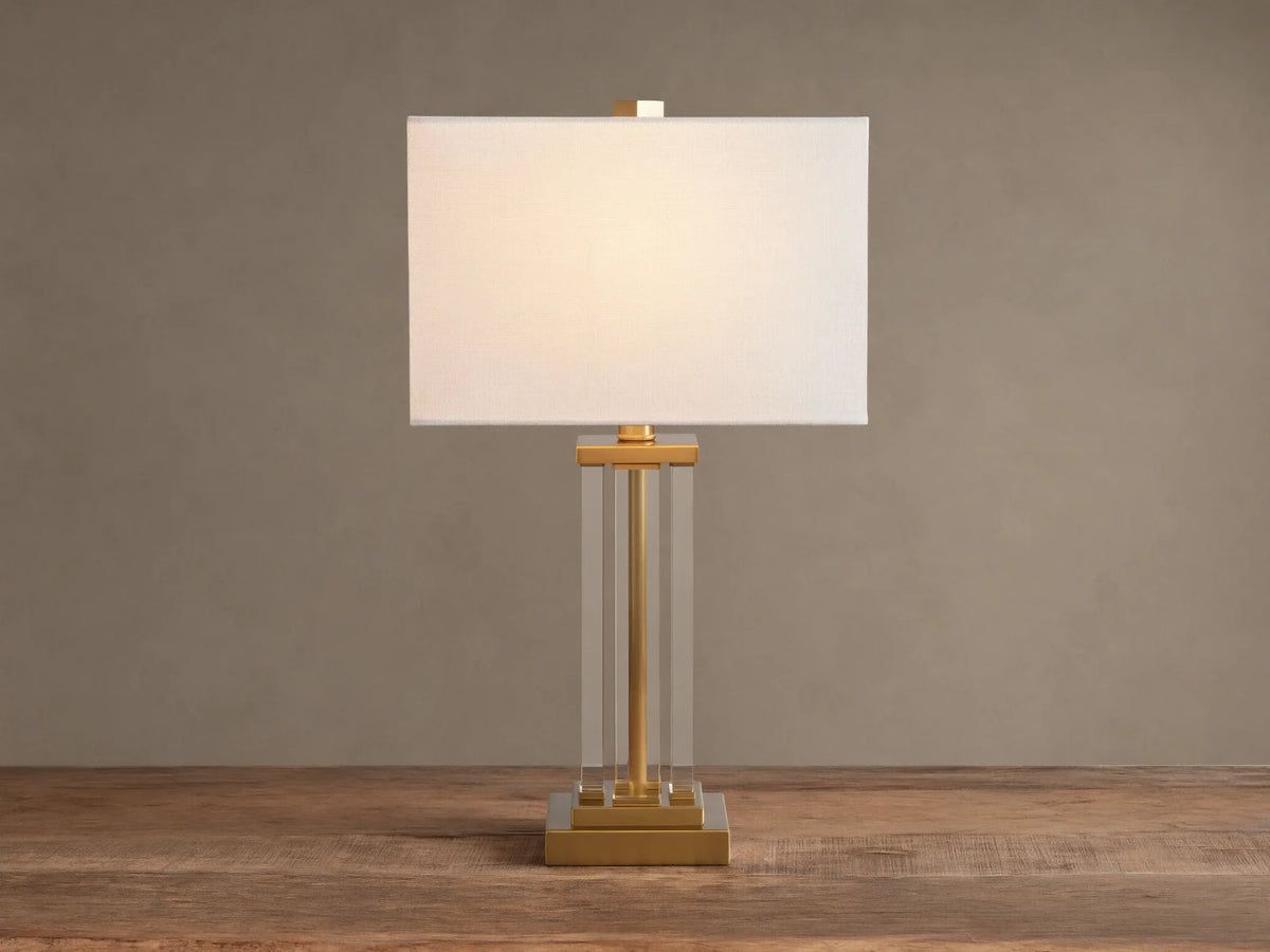 Dawson Table Lamp: white rectangular shade, clear acrylic column body, and gold-tone square base on a wood surface.