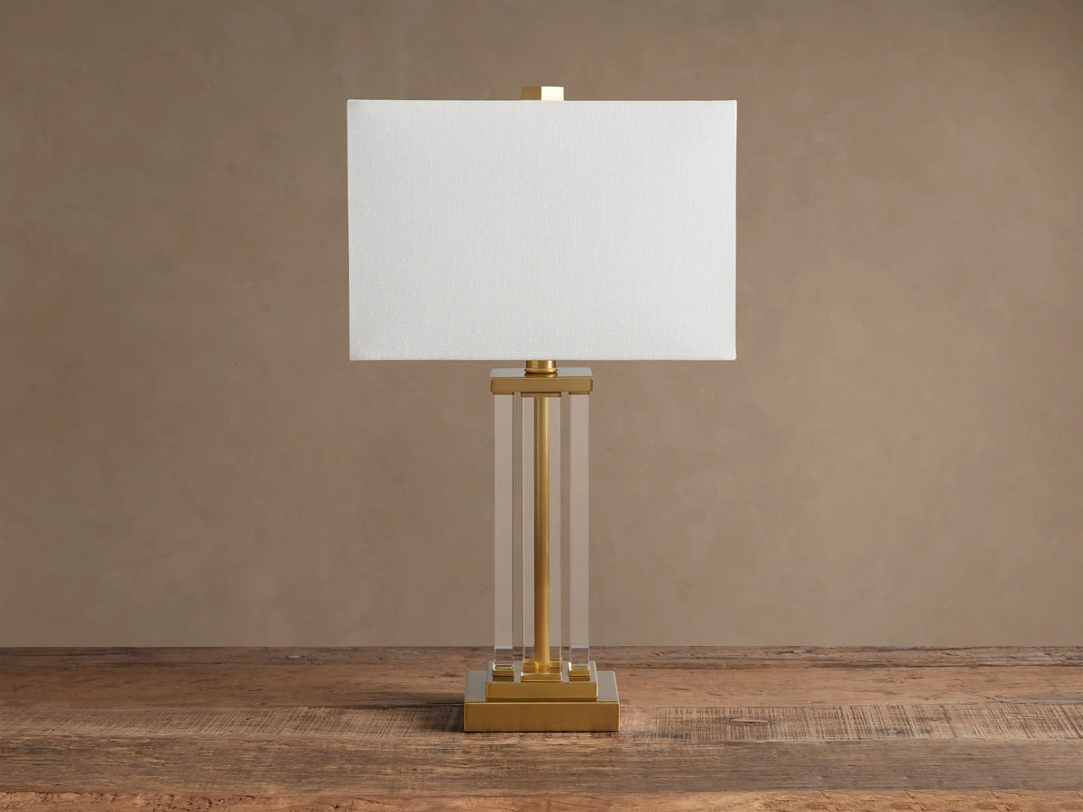 Dawson Table Lamp: white rectangular shade, clear acrylic column body, and gold-tone square base on a wood surface.
