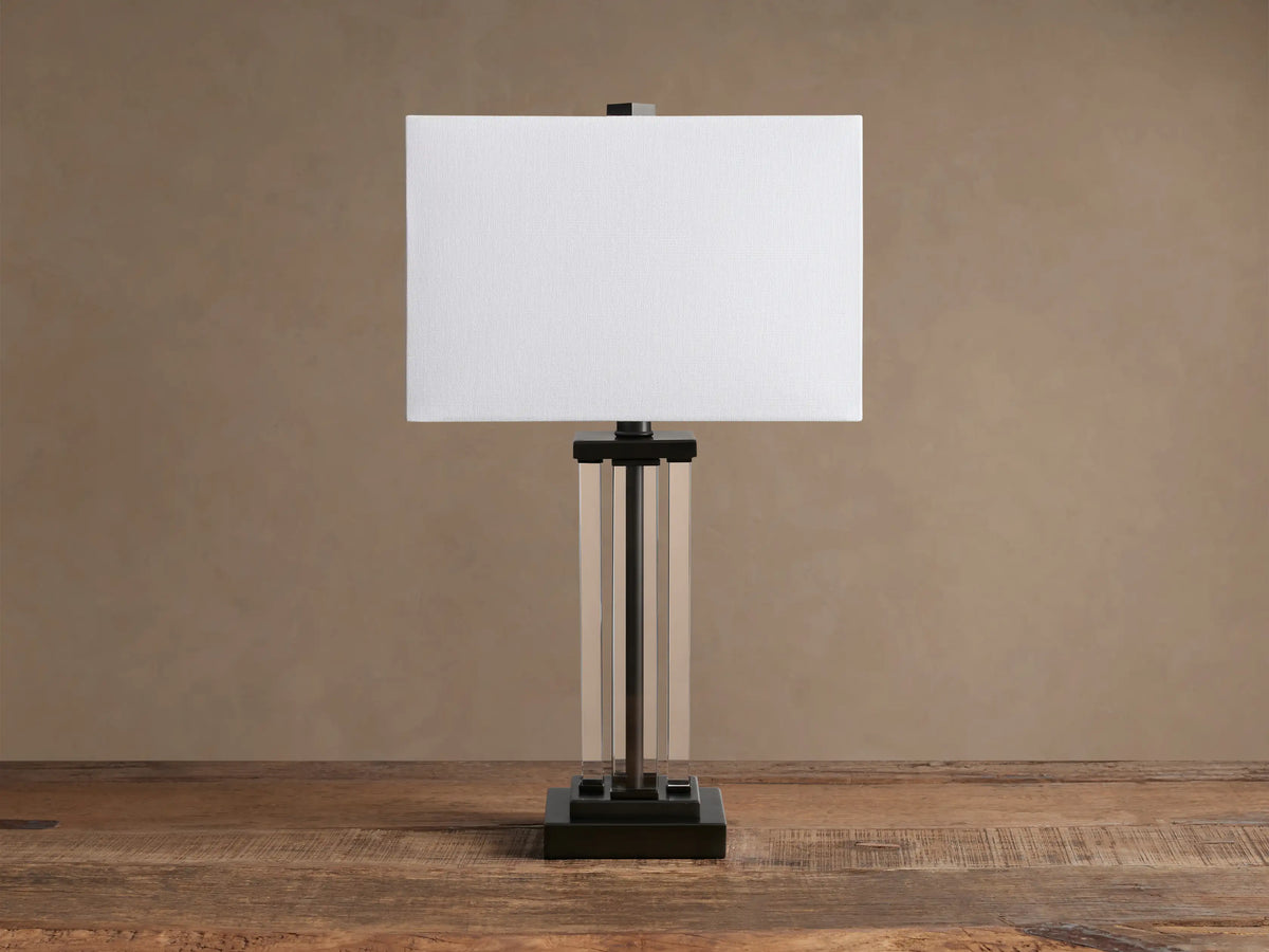 Dawson Table Lamp with a lit rectangular white fabric shade, clear acrylic pillars, and a dark bronze square base on a wooden table.