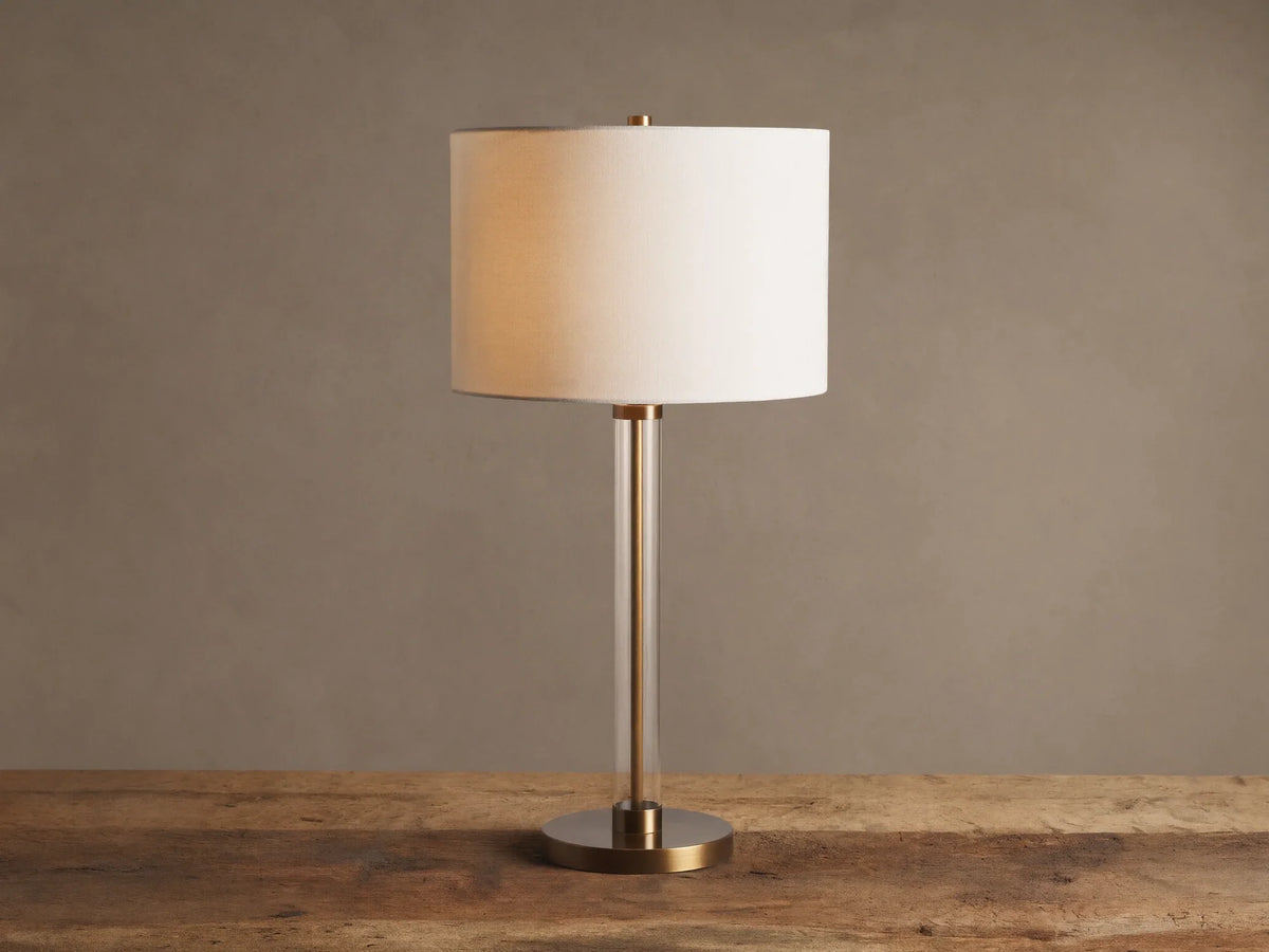 Lit Glass Column Table Lamp featuring a clear glass stem, bronze base, and cream linen drum shade on a rustic wood table.