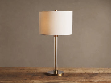 Lit Glass Column Table Lamp featuring a clear glass stem, bronze base, and cream linen drum shade on a rustic wood table.