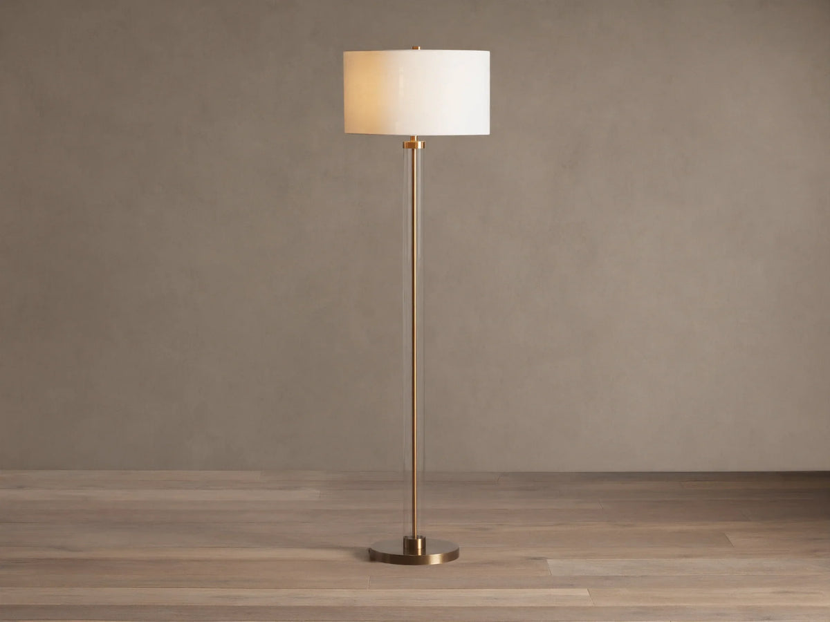 Clear glass column floor lamp with a brass base and stem, and a cylindrical cream linen shade. Standing on a wooden floor.