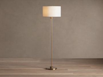 Clear glass column floor lamp with a brass base and stem, and a cylindrical cream linen shade. Standing on a wooden floor.