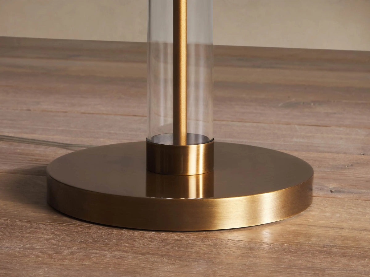 Tall Glass Column Floor Lamp with clear glass column, brass base, and a white cylindrical linen shade, standing on a wood floor.