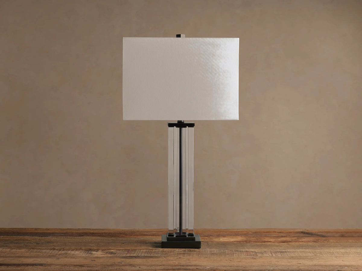 Dawson Table Lamp with glowing white rectangular shade, clear and dark metal base, on a rustic wooden table.