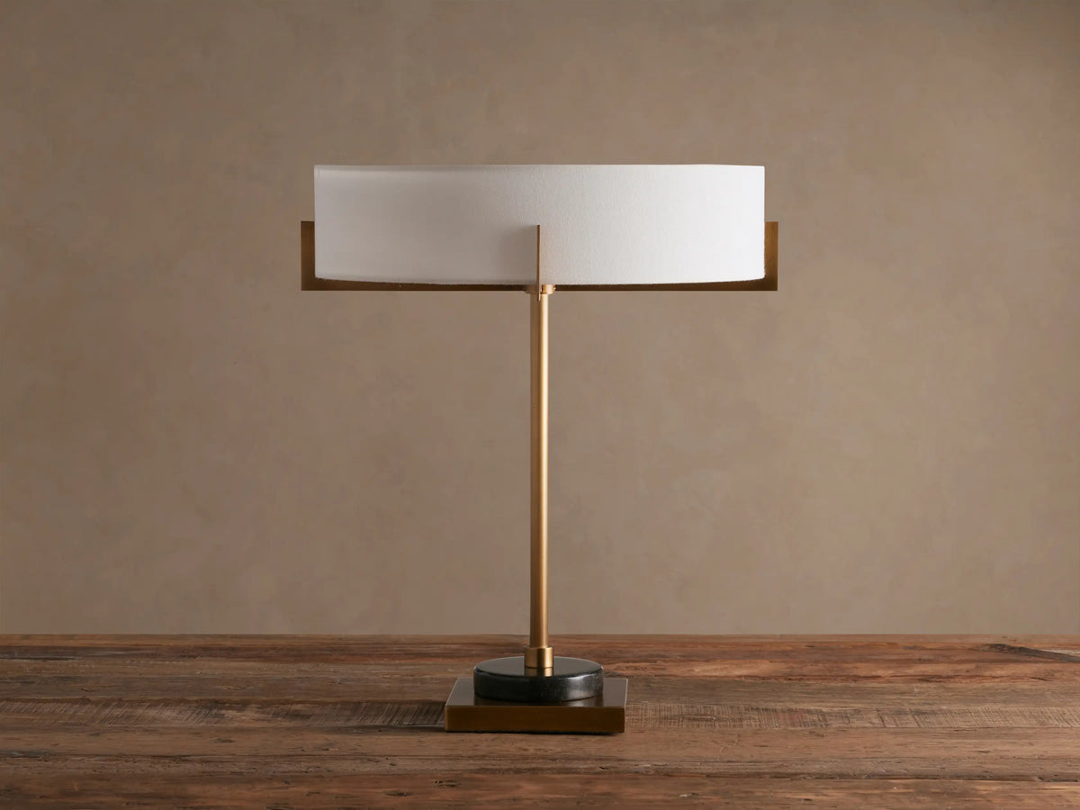 Frederic Table Lamp with antiqued brass finish, black granite base, and illuminated white linen shade on a wooden table.