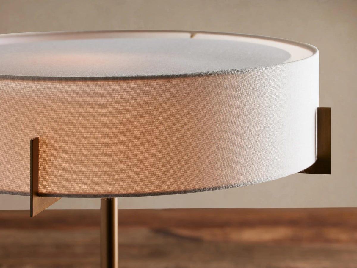 Frederic Table Lamp with antiqued brass frame, black granite base, and cylindrical white linen shade, on a wooden table.