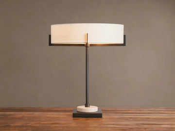 Illuminated Frederic Table Lamp with a bronze frame, white linen shade, and a cream marble disc on a dark square base, on wood.